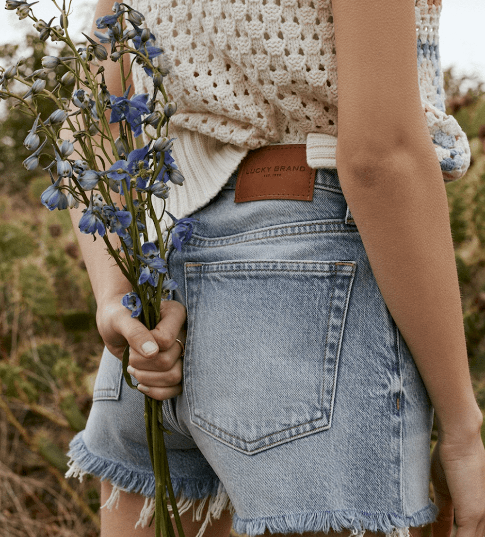 close up image of a woman in jean shorts and a knit crochet top holding flowers behind her back