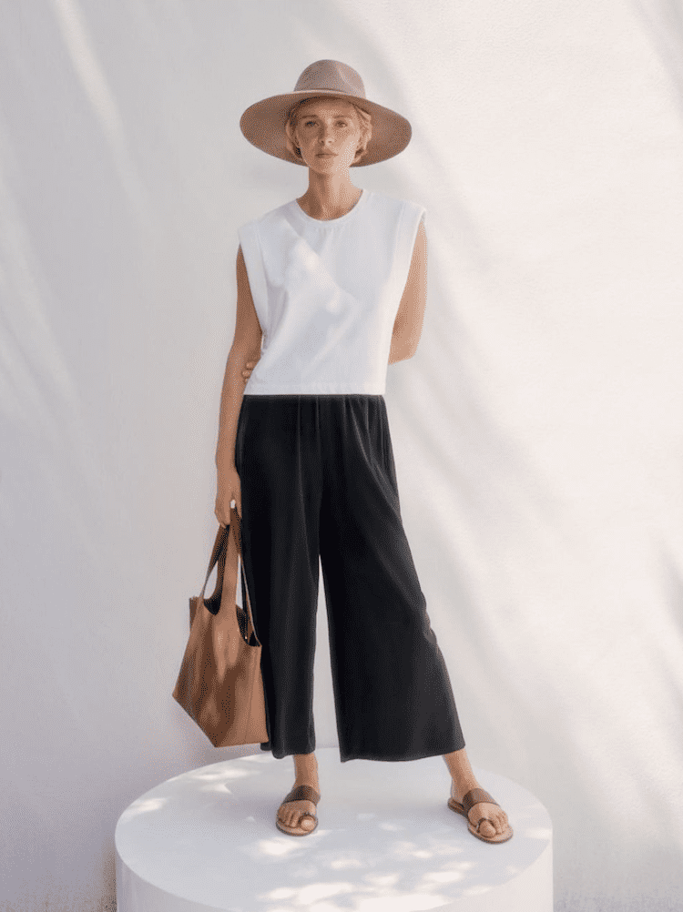image of a woman standing on a white platform wearing a white tank top, black wide leg pants, sandals, and a sun hat