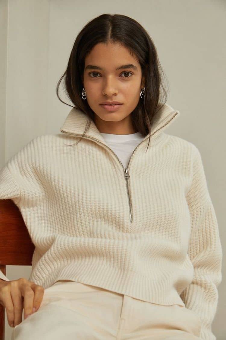 image of a woman with brown hair sitting back in a chair wearing a knit half zip cream sweater and cream jeans