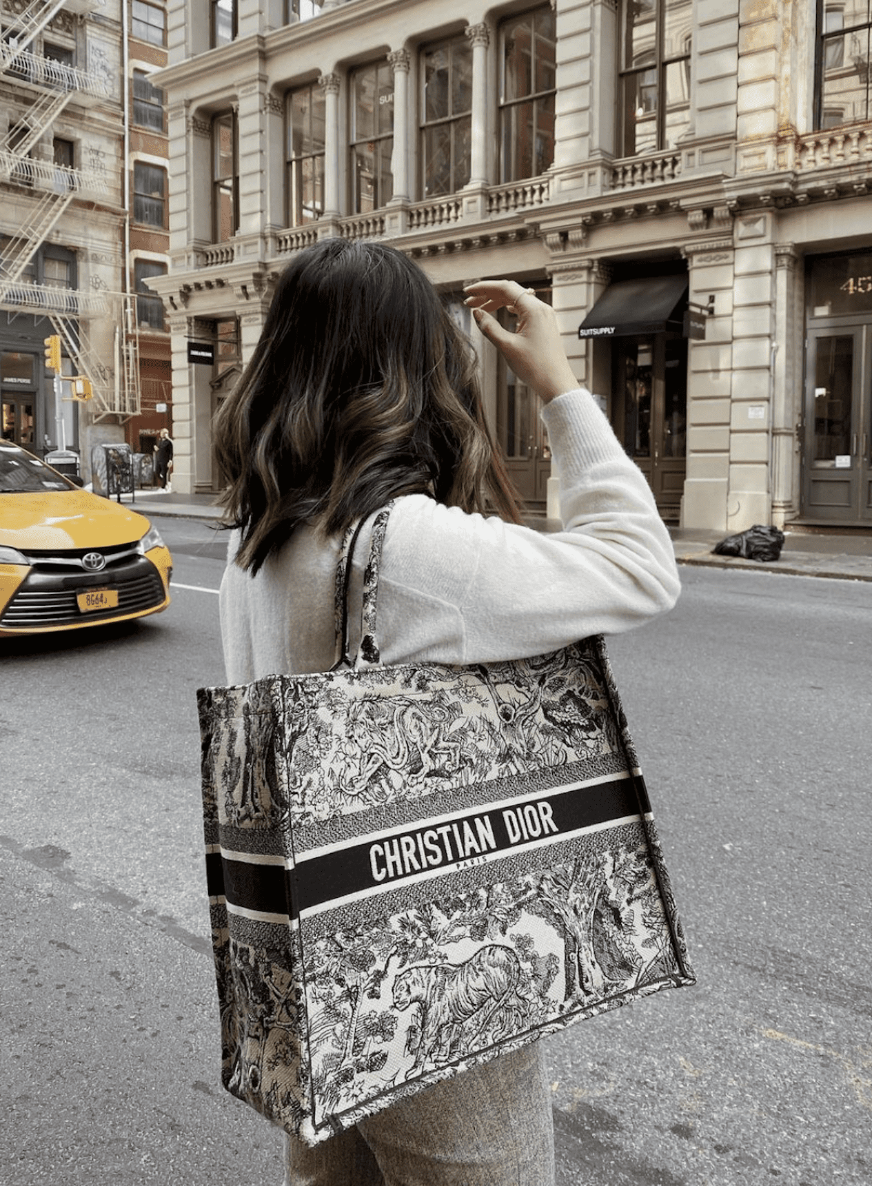 image of a woman wearing a cream sweater with a Dior book tote bag standing on a busy street with her head turned to the side