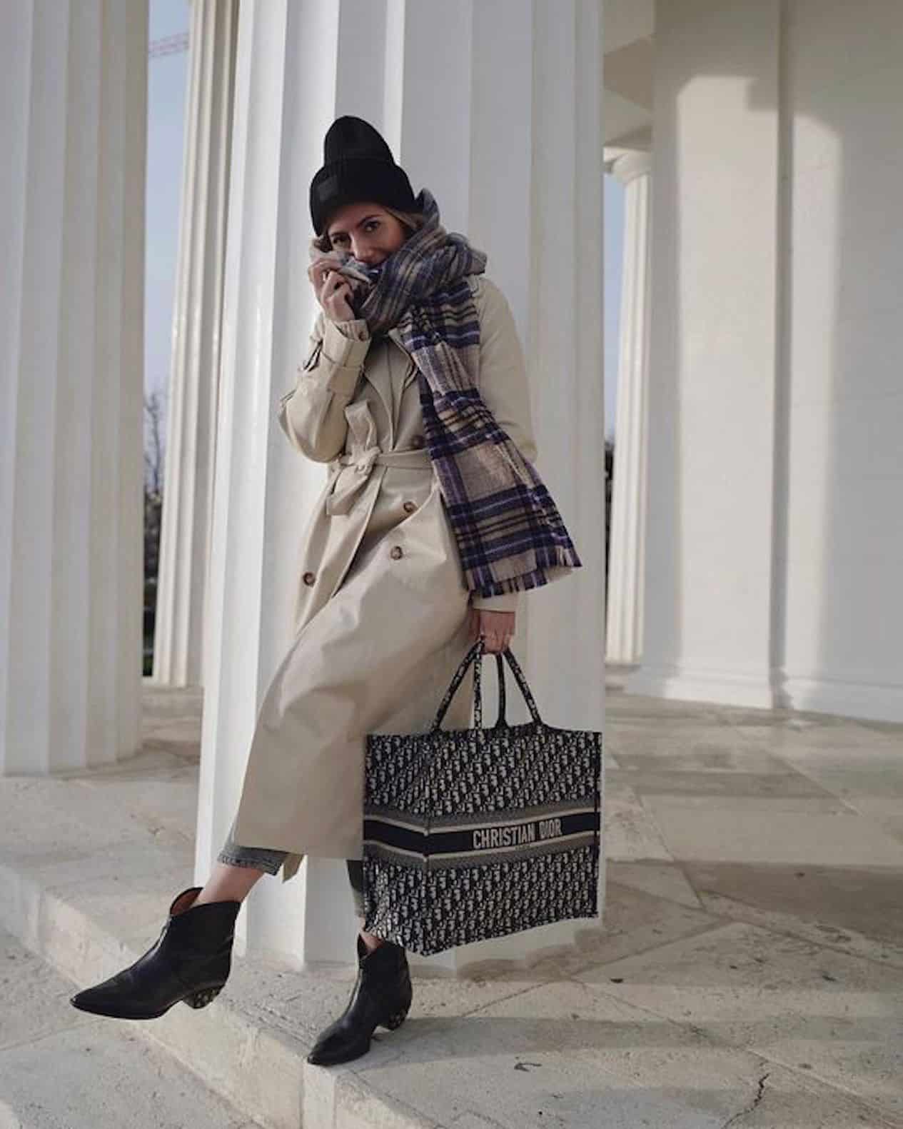 image of a woman standing against a large column wearing black boots, a beige trench coat, a big scarf, a toque, and holding a Dior book tote bag