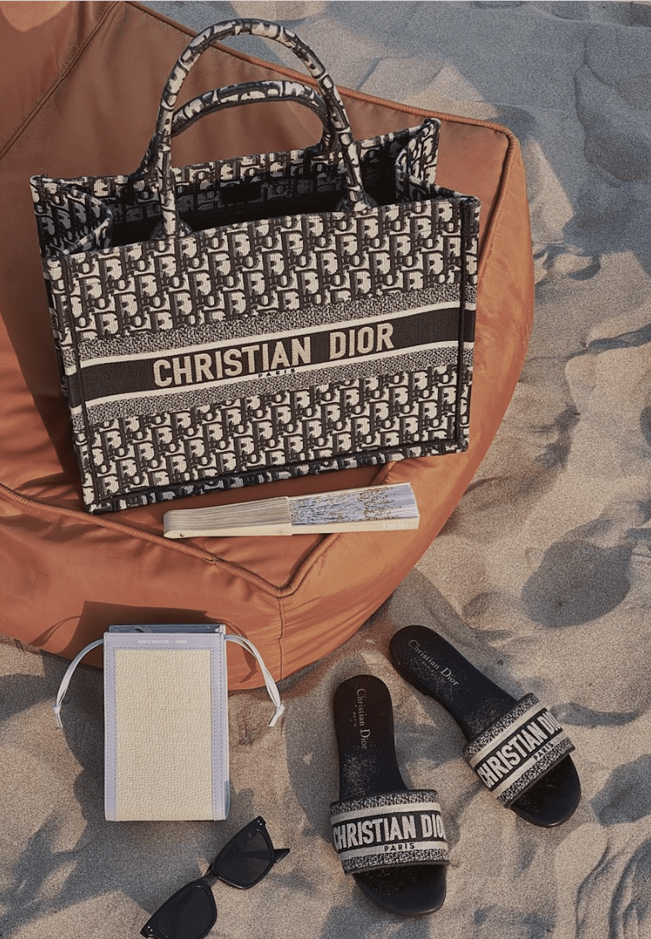 image of a Dior book tote back on a pouf on the beach with Dior sandals and sunglasses beside
