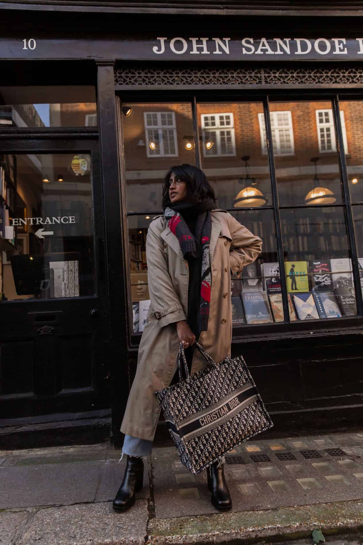 image of a brown woman standing in front of a book store wearing a long tan trench coat and holding a Dior book tote