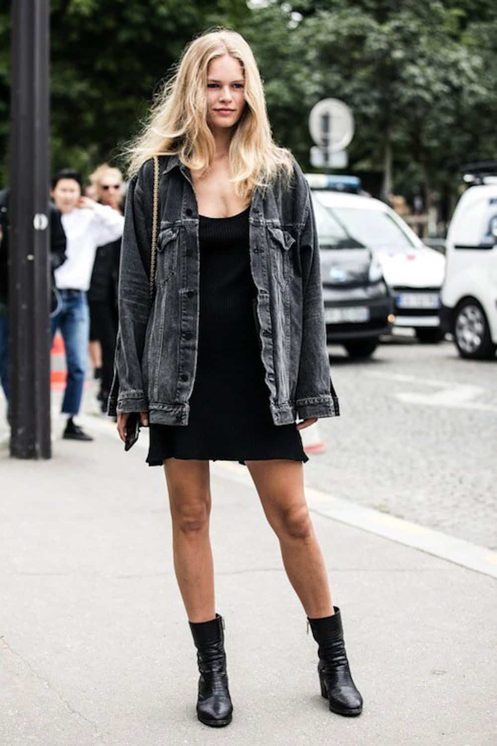 image of a woman standing on the sidewalk wearing an oversized black jean jacket, a black mini dress, and black ankle boots