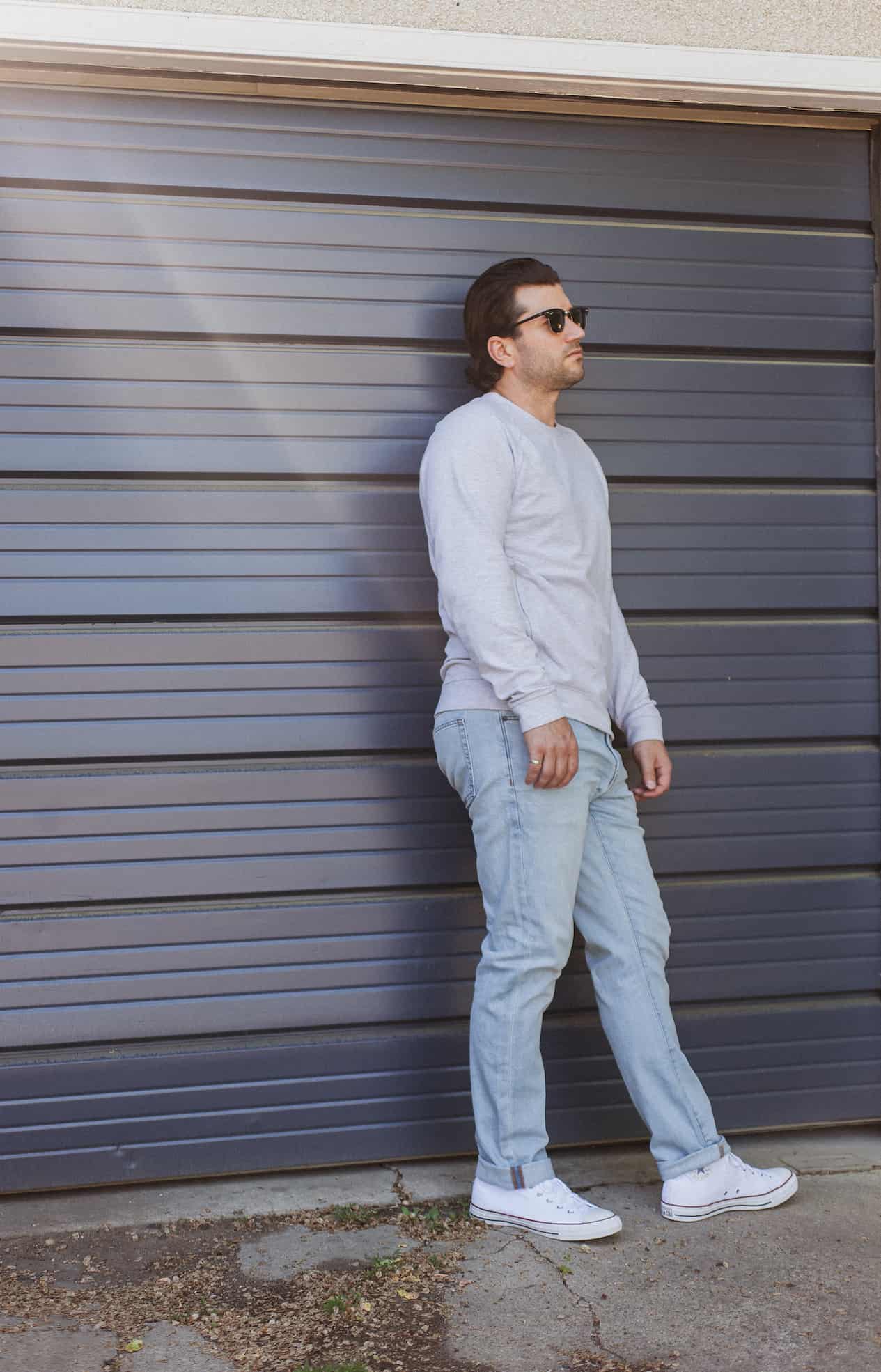 image of a man with brown hair standing against a dark blue garage door wearing a grey sweater, light blue jeans, and white sneakers