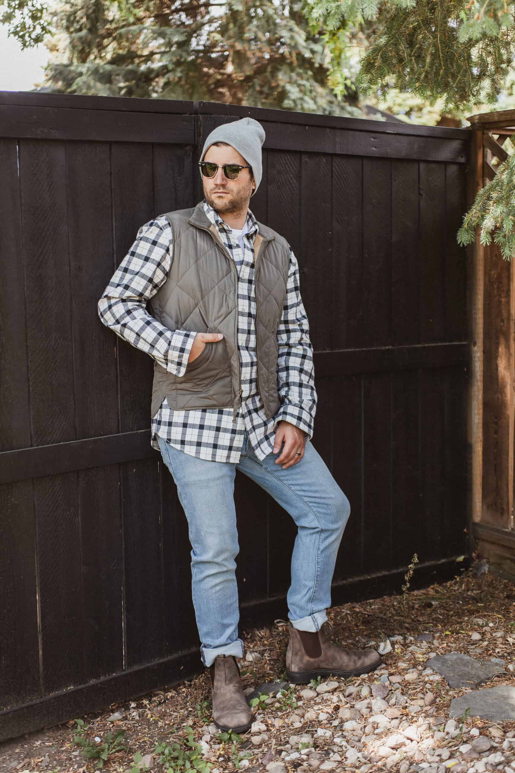 image of a man leaning against a black fence wearing a white and black flannel shirt, green vest, light blue jeans, and Blundstone boots
