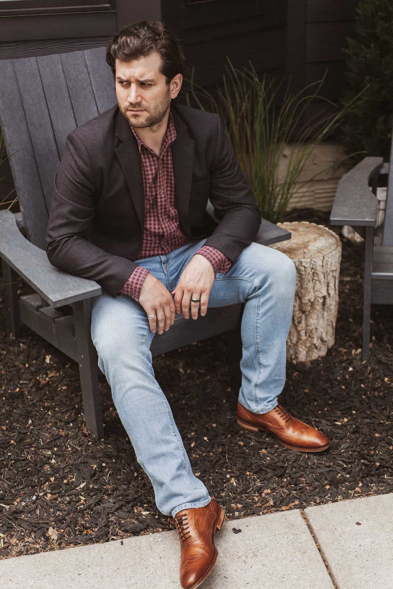 image of a man sitting on a black Adirondack chair wearing a charcoal blazer, red plaid shirt, light blue jeans, and brown shoes