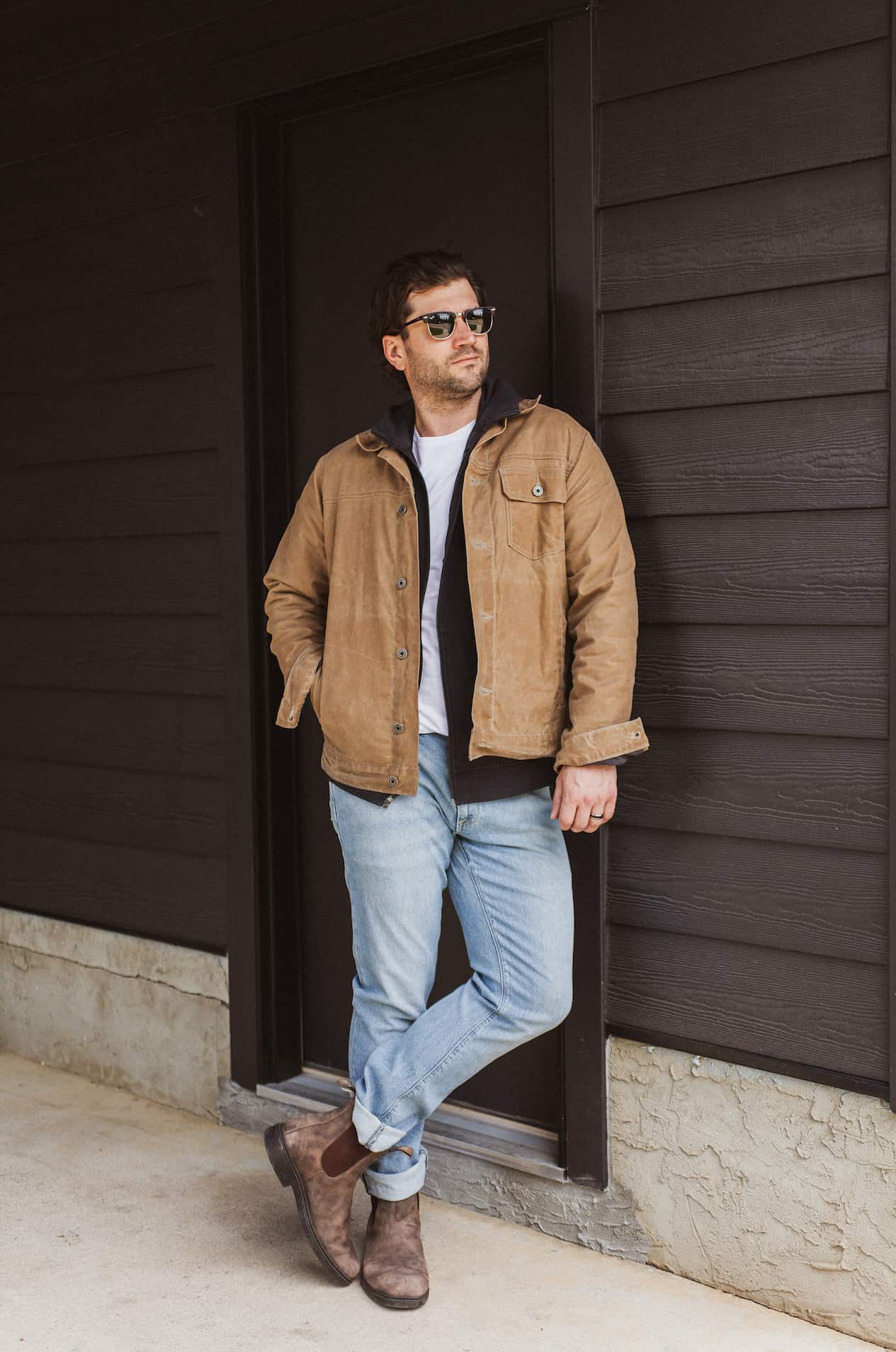 image of a man standing against a black wall wearing a brown trucker jacket, black sweater, light blue jeans, and blundstone boots