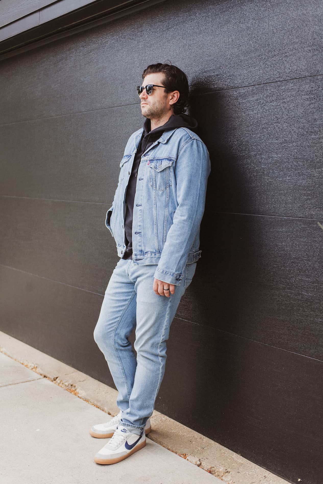 image of a man leaning against a black garage door wearing a denim jacket, black hoodie, light blue jeans, and white sneakers