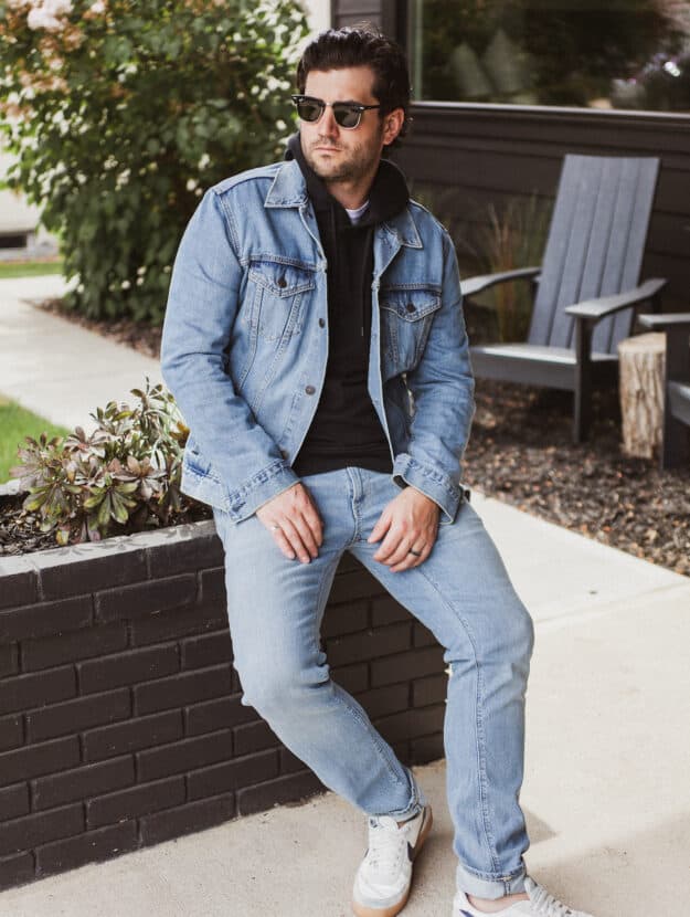 image of a man sitting on a brick planter wearing a denim jacket, black hoodie, light blue jeans, and white sneakers
