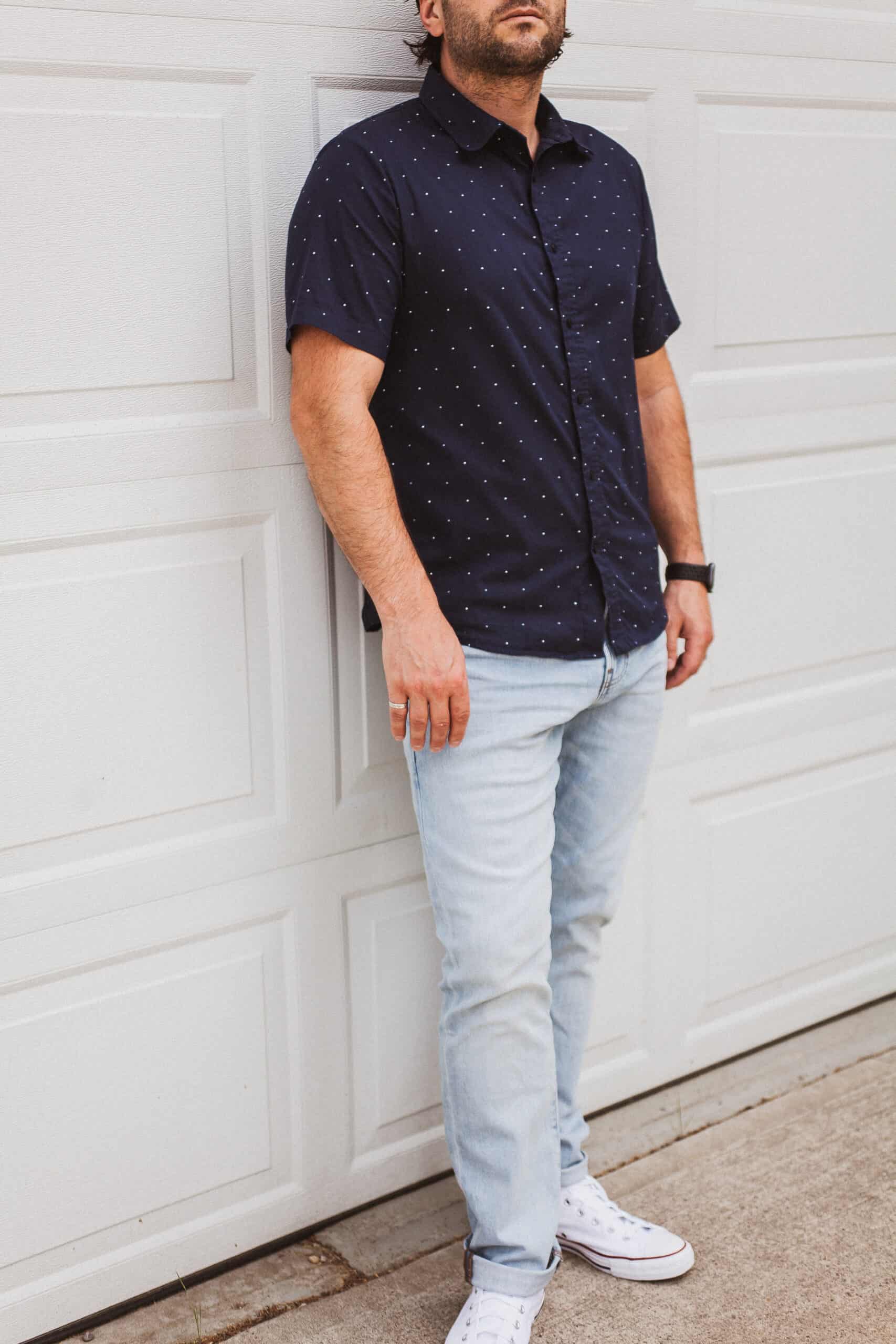 image of a man leaning against a white garage door wearing a navy blue button-up short sleeve shirt, light blue jeans, and white sneakers 