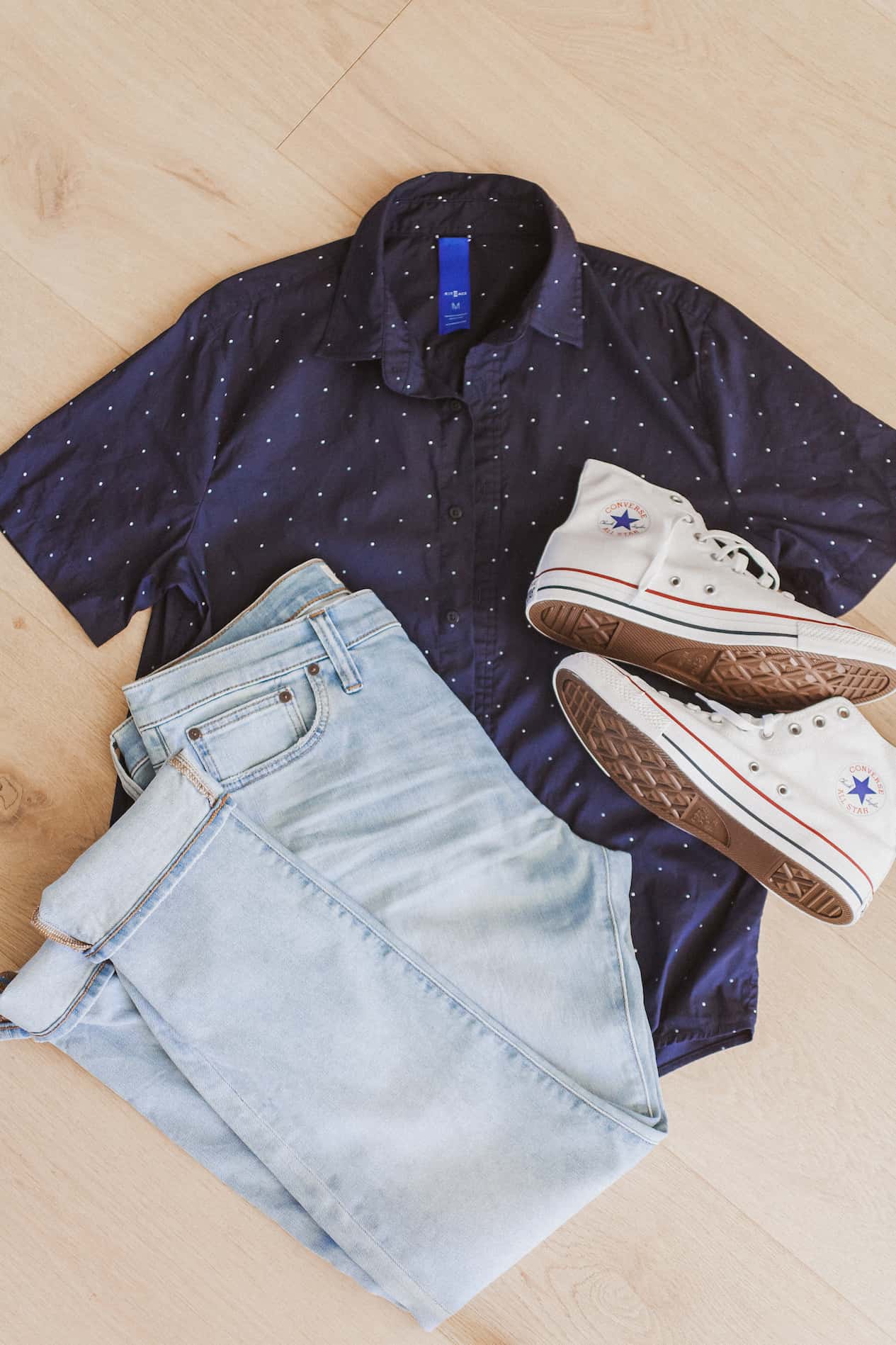 image of an outfit flat lay with a navy blue polka dot button up t-shirt, light blue jeans, and white Converse high top sneakers