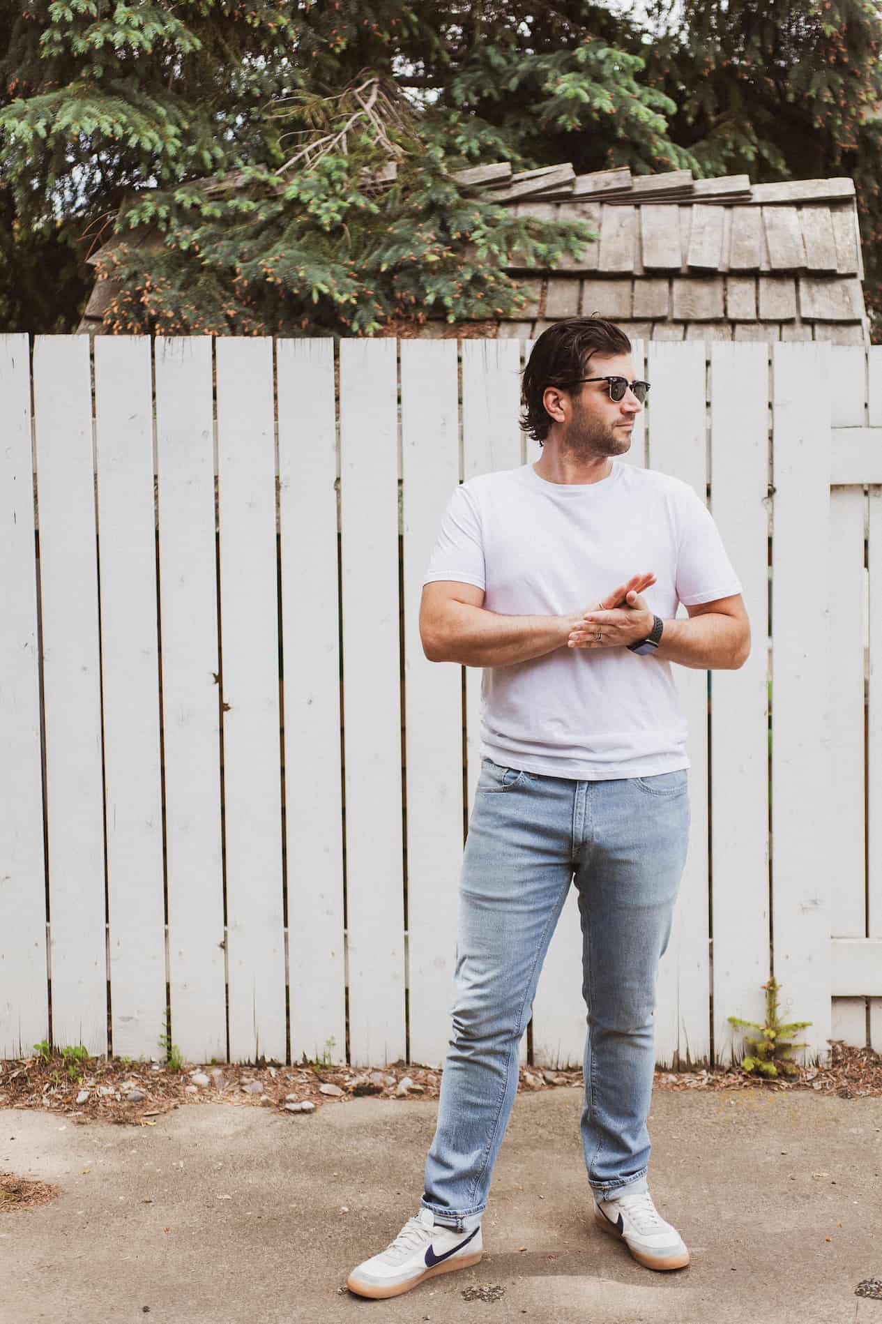 image of a man standing in front of a white fence wearing a white t-shirt and light blue jeans with white sneakers