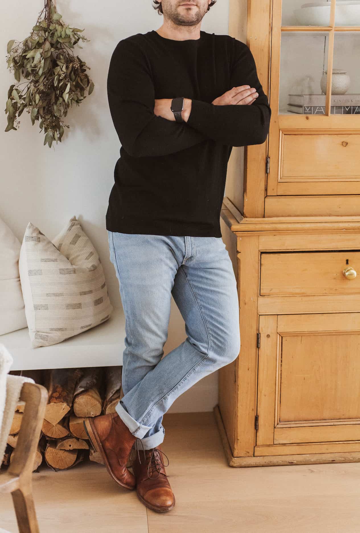 image of a man from the neck down standing against a wood cabinet wearing a black sweater, light blue jeans, and brown boots