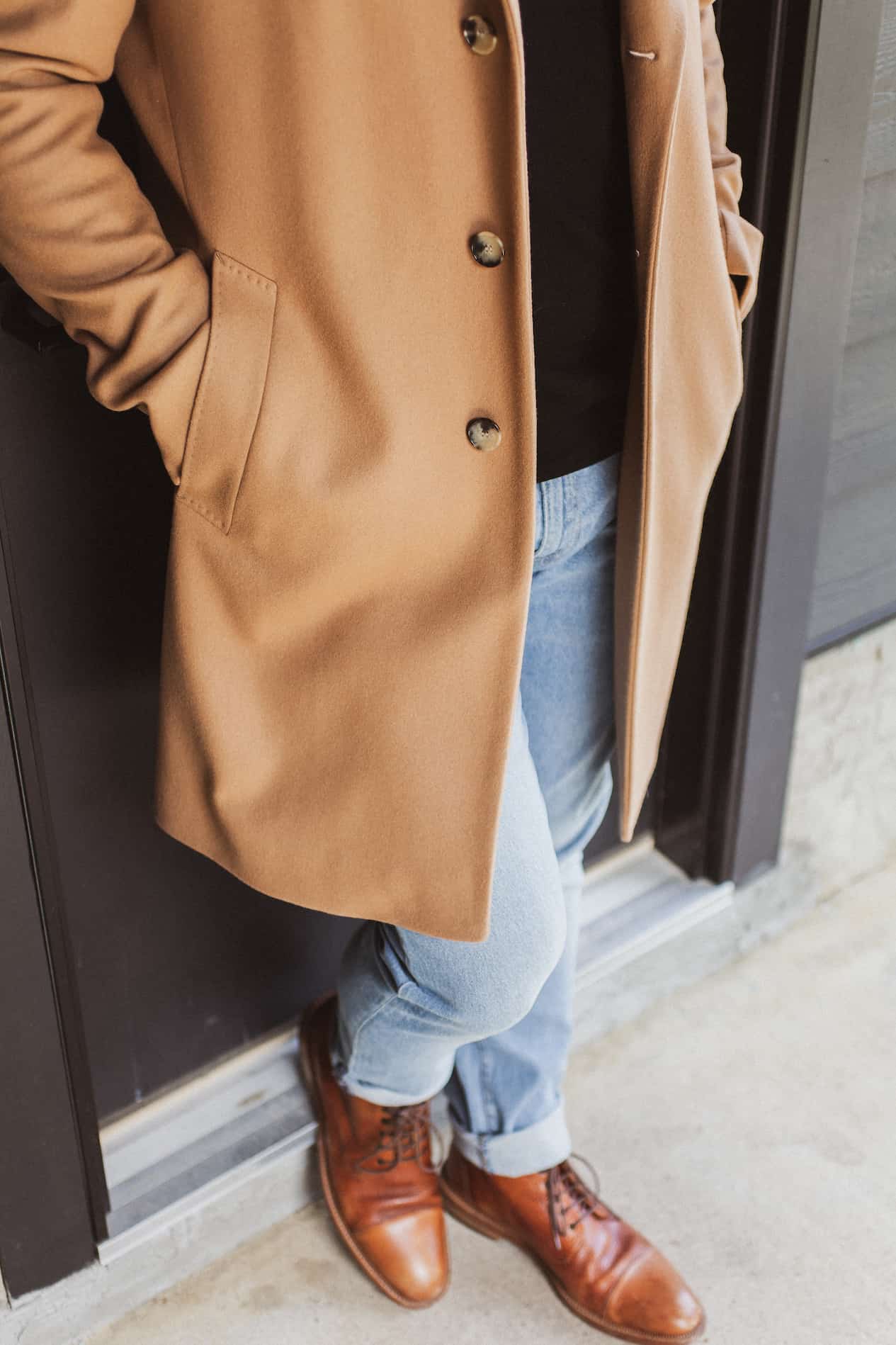 image of a man from the shoulders down wearing a long wool camel coat, black shirt, light blue jeans, and brown boots