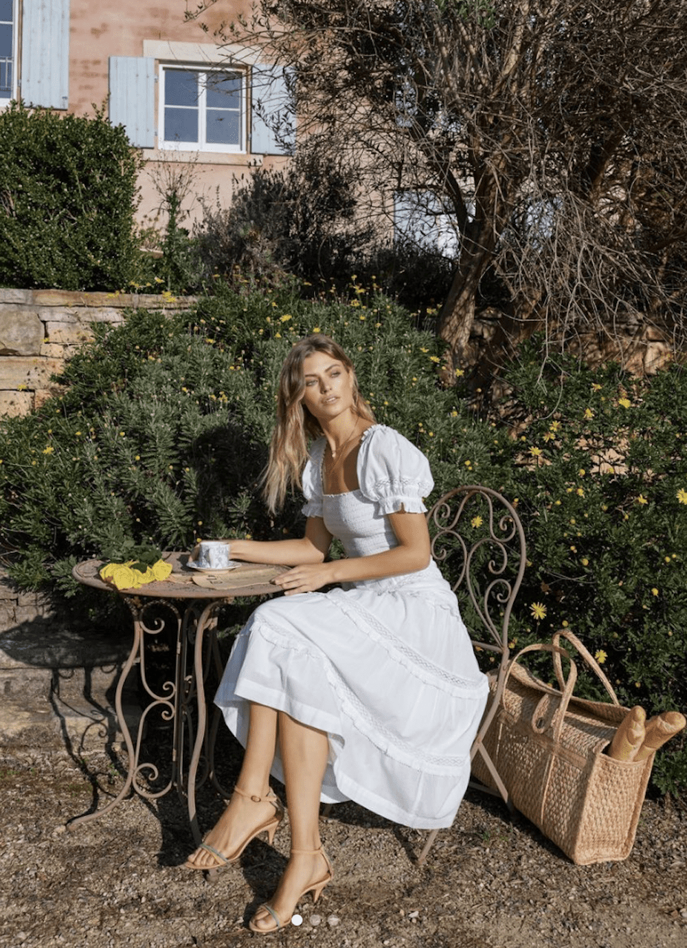 image of a blonde woman sitting at a cafe table outside in a garden wearing a pretty white dress