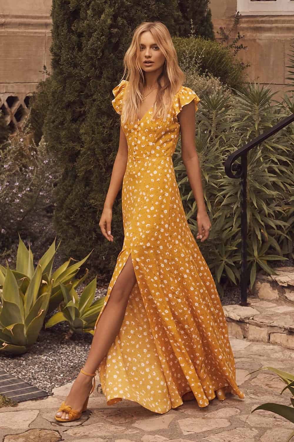 image of a woman wearing a long yellow floral maxi dress walking down a sidewalk with green plants in the background 