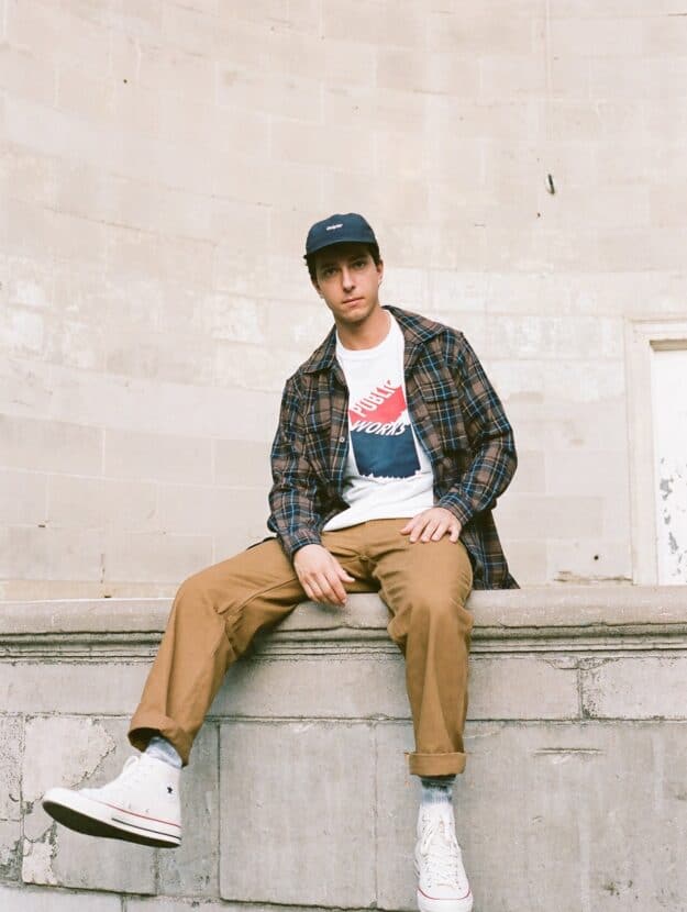 image of a man sitting on a concrete ledge wearing khaki pants, sneakers, a white graphic t-shirt, and a plaid shirt over top