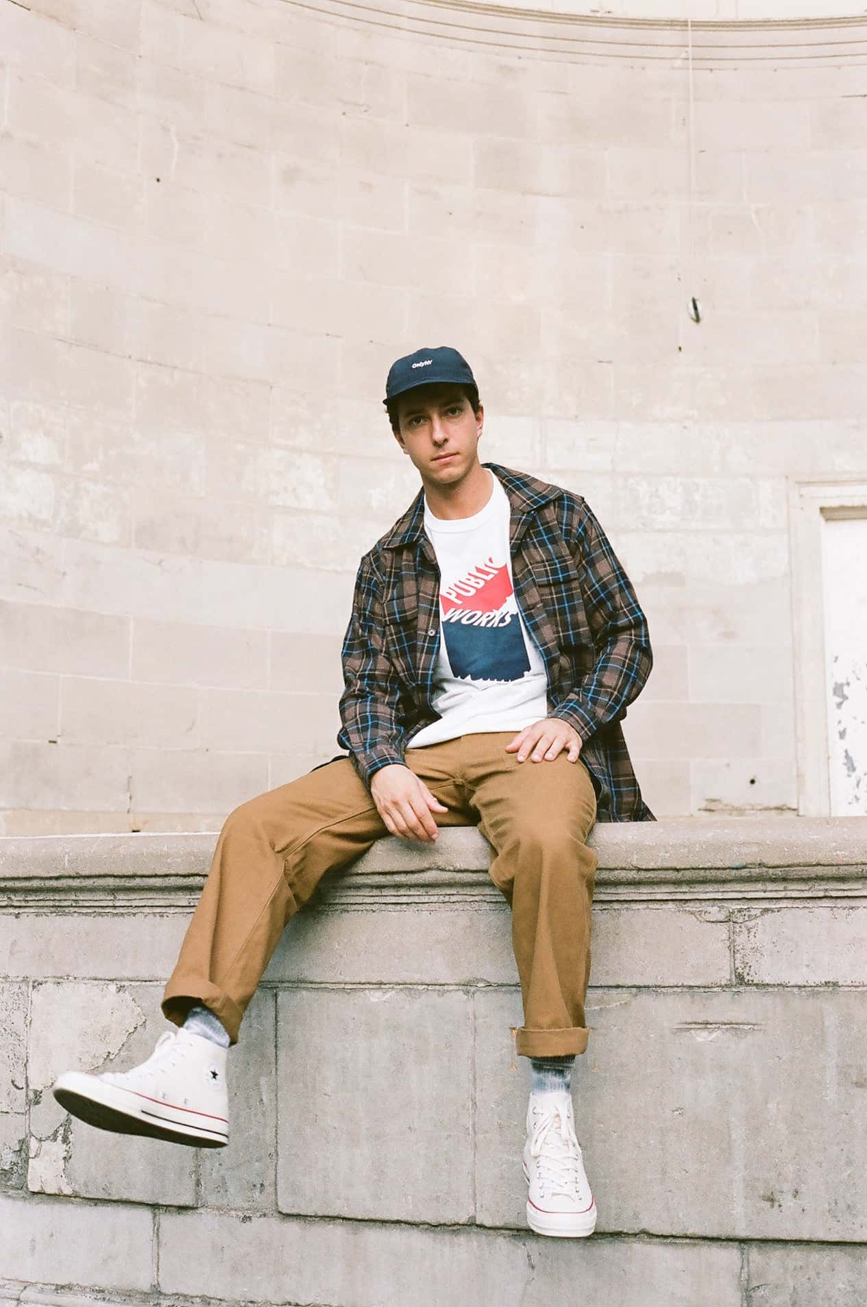 image of a man sitting on a concrete ledge wearing khaki pants, sneakers, a white graphic t-shirt, and a plaid shirt over top 