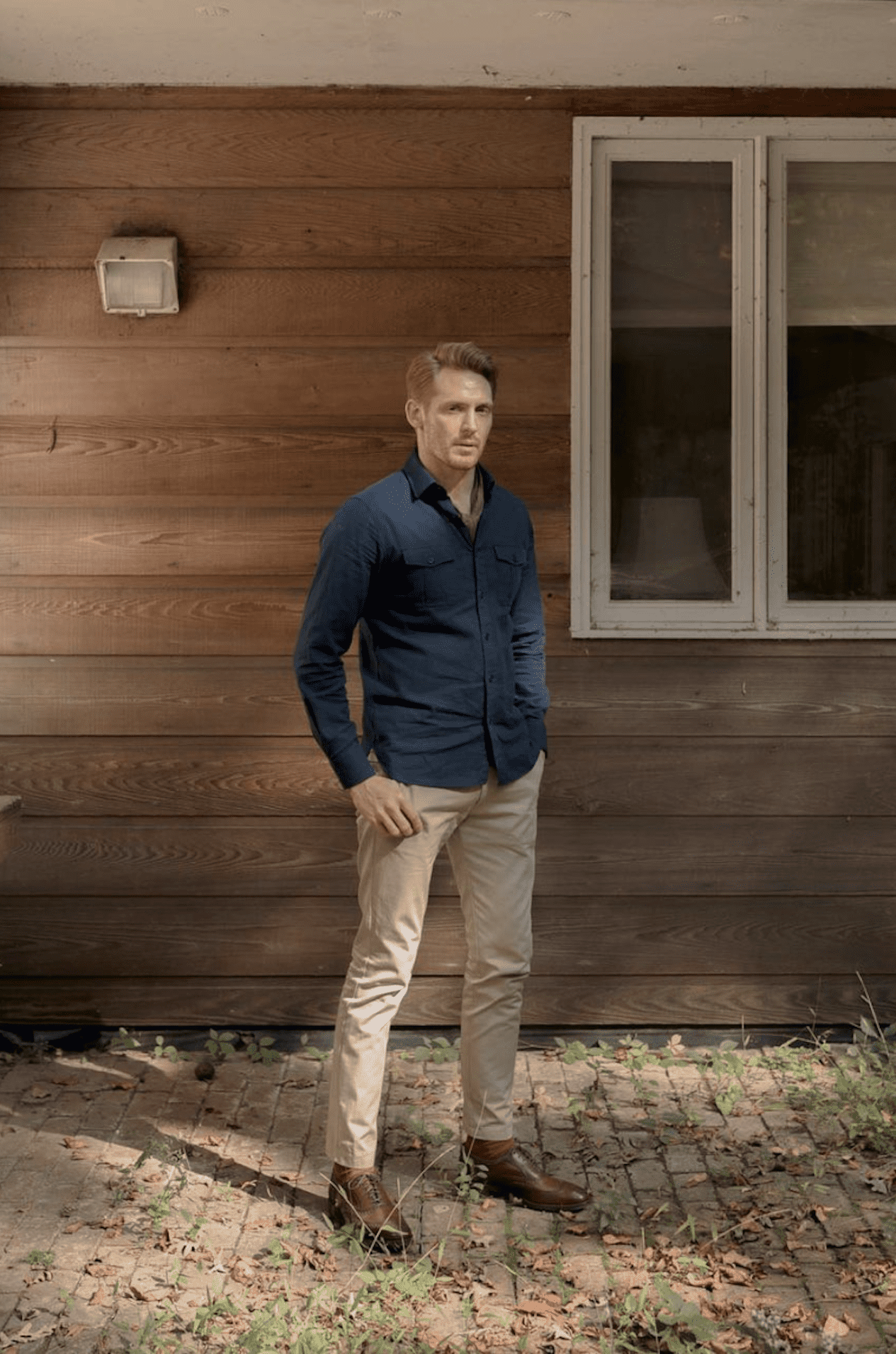 image of a man standing in front of a brown house wearing a blue button up shirt, khaki pants, and brown boots