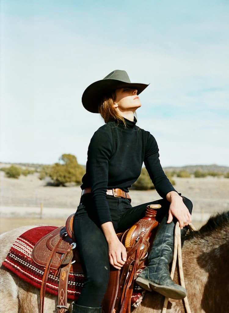 image of a woman wearing a black long sleeve shirt, western buckle belt, cowboy hat, and black jeans sitting on a horse