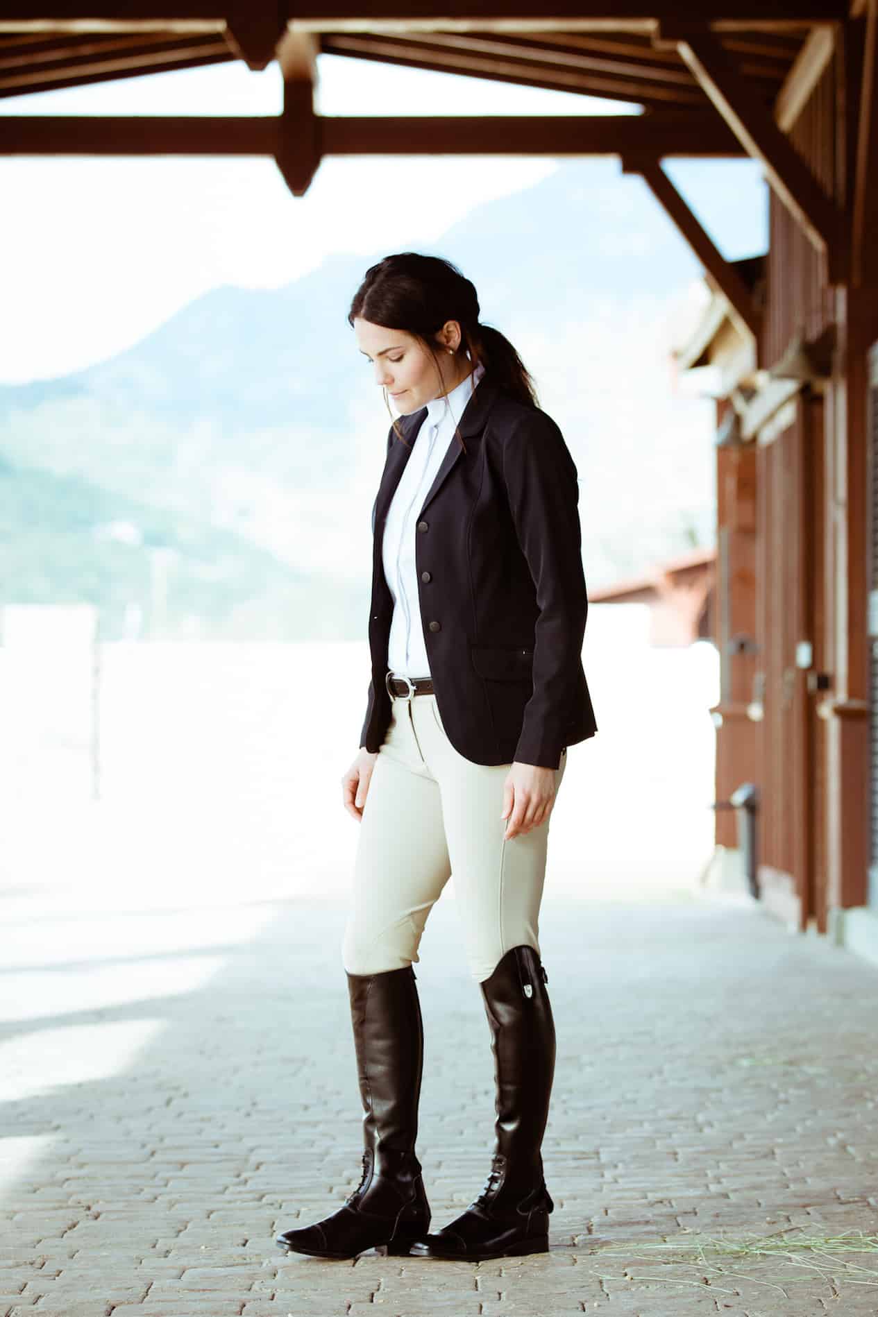 image of a woman standing in stables wearing an English equestrian outfit with a blazer, white shirt, riding leggings, and riding boots