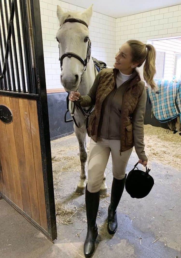 image of a woman standing in a horse stable with a horse wearing riding leggings, riding boots, a sweater, and a vest