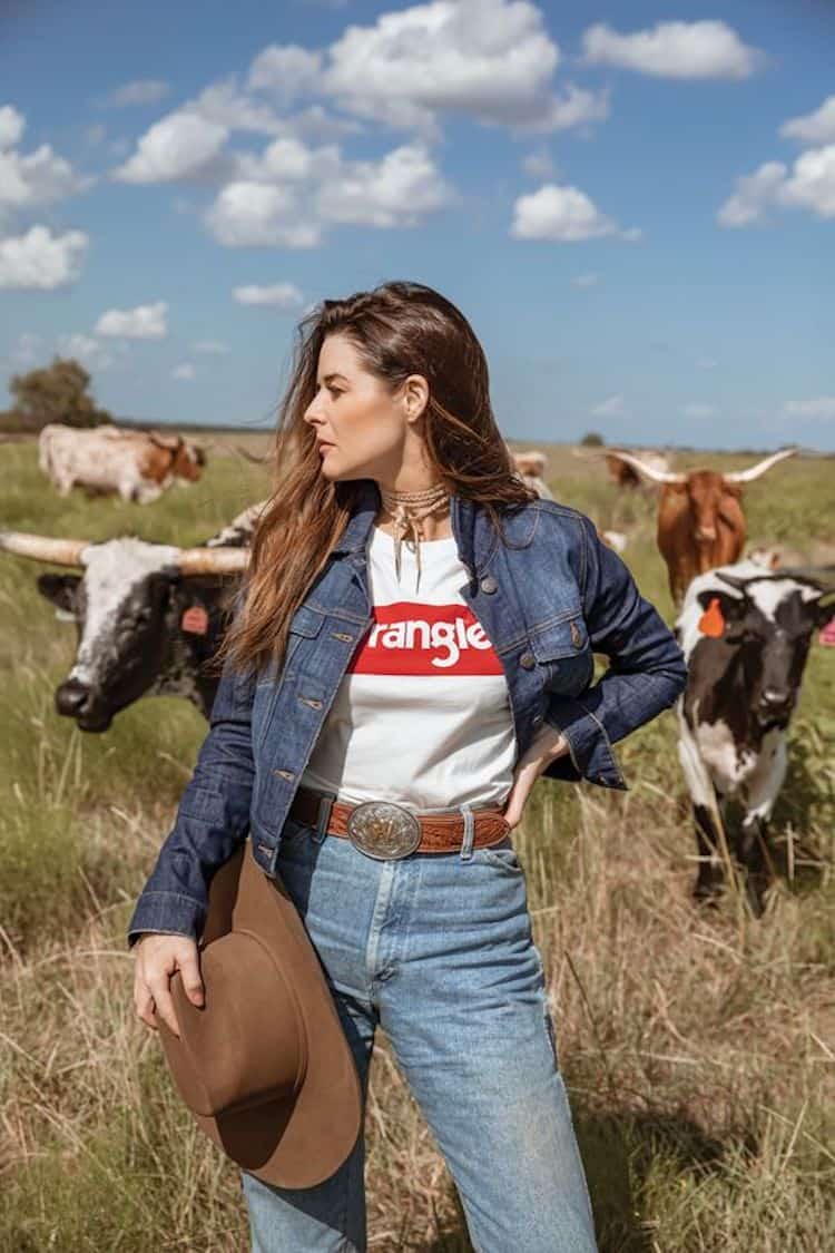 image of a woman standing in a field of cows wearing a wrangler t-shirt, denim button up, jeans, and a belt with large buckle