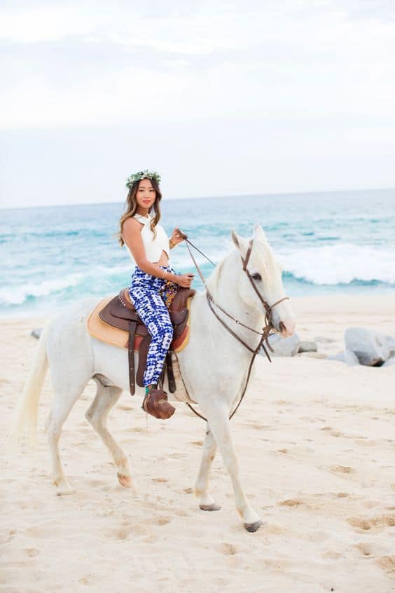 image of a woman on the beach horseback riding in blue and white pants and a white crop top