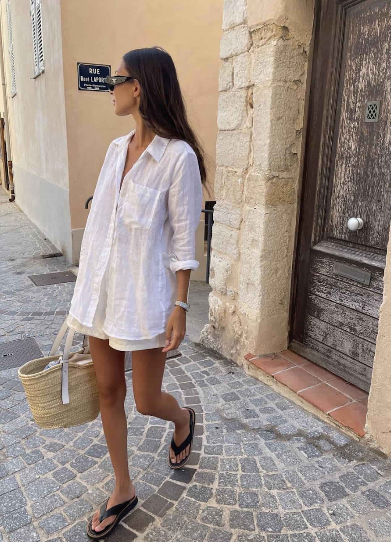image of a woman walking down a road wearing a white linen button up shirt, shorts, and sandals