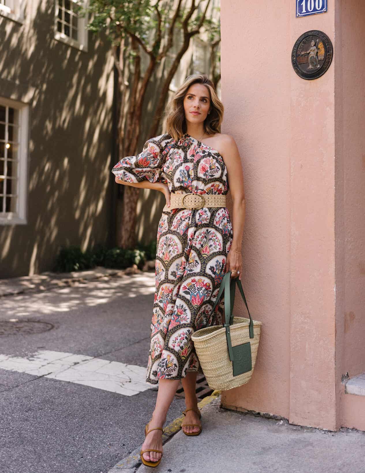 image of a woman wearing a bright patterned maxi one shoulder dress, holding a straw tote, and wearing sandals