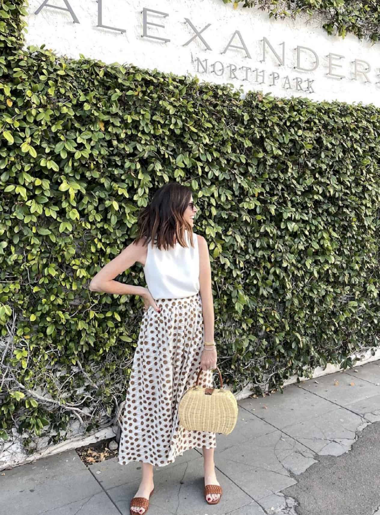 image of a woman standing in front of a wall with vines wearing a polka dot midi skirt and white blouse