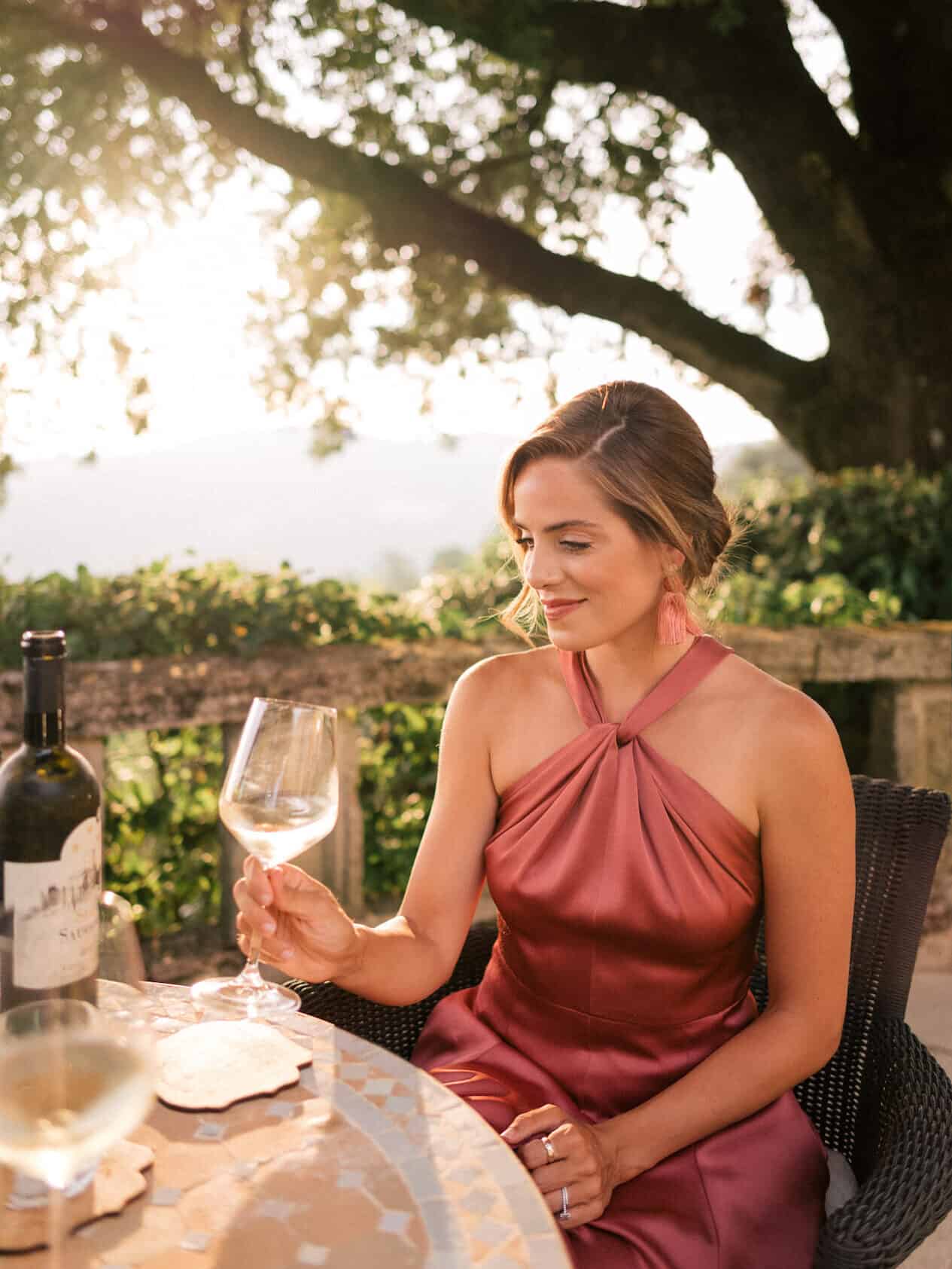 image of a woman in a dark blush satin dress sitting at a table drinking a glass of wine