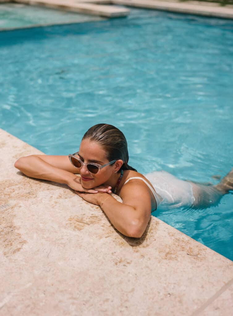 image of a woman in a swimming pool leaning on the edge of the pool wearing a white swimsuit and sunglasses