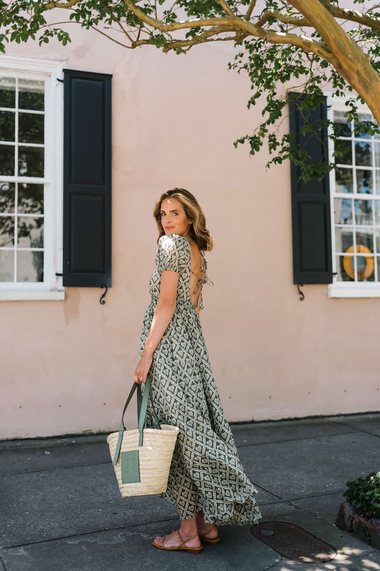 image of a woman standing on a sidewalk in front of a pink wall wearing a green maxi floral dress holding a woven tote bag