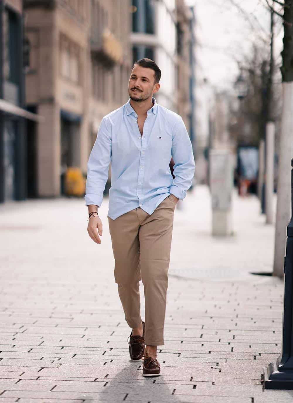 image of a man wearing brown shoes, khaki pants, and a light blue button up oxford shirt, as he walks down the sidewalk