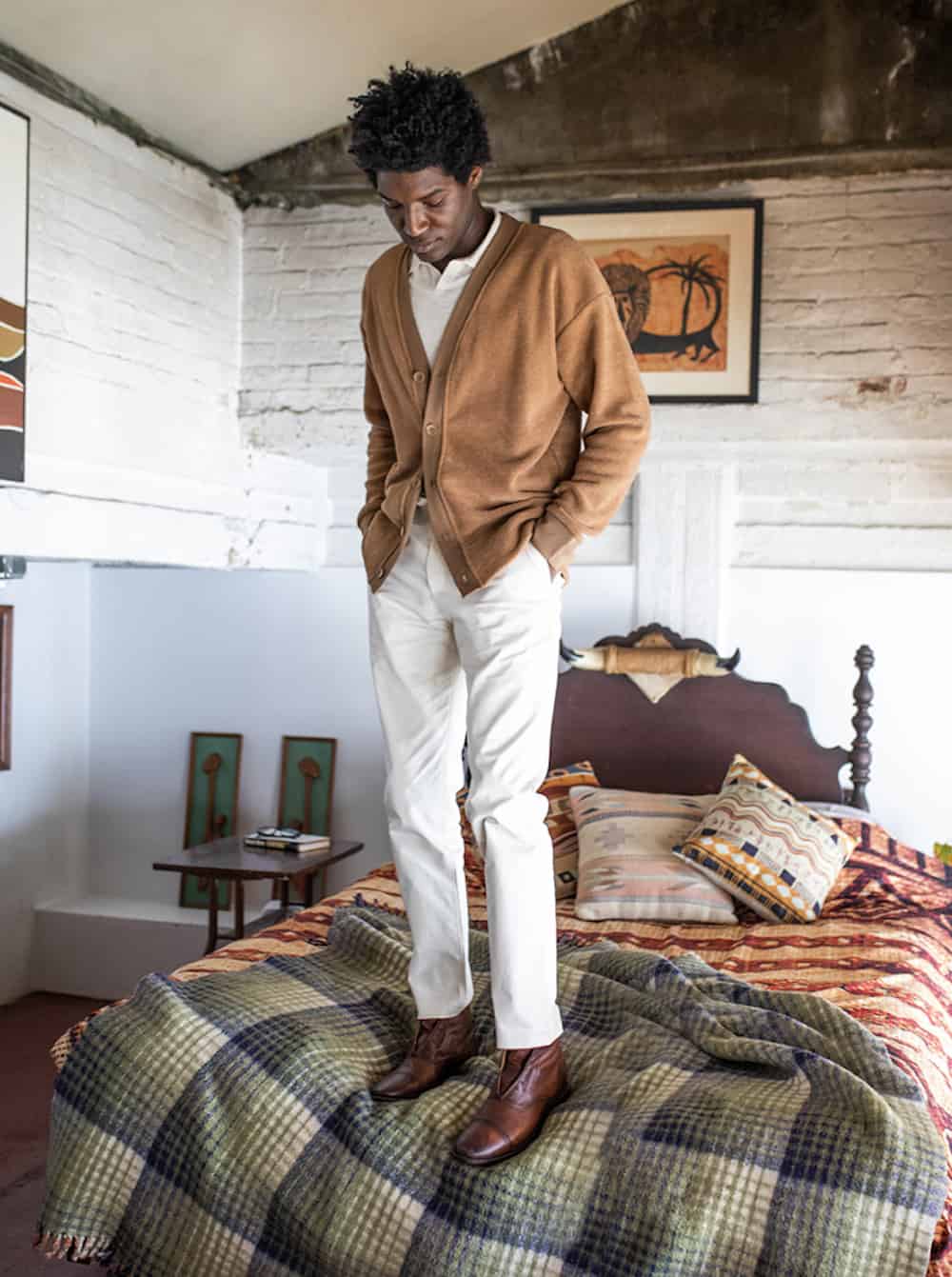 image of a black man standing on a bed wearing brown shoes, khaki pants, and a brown cardigan