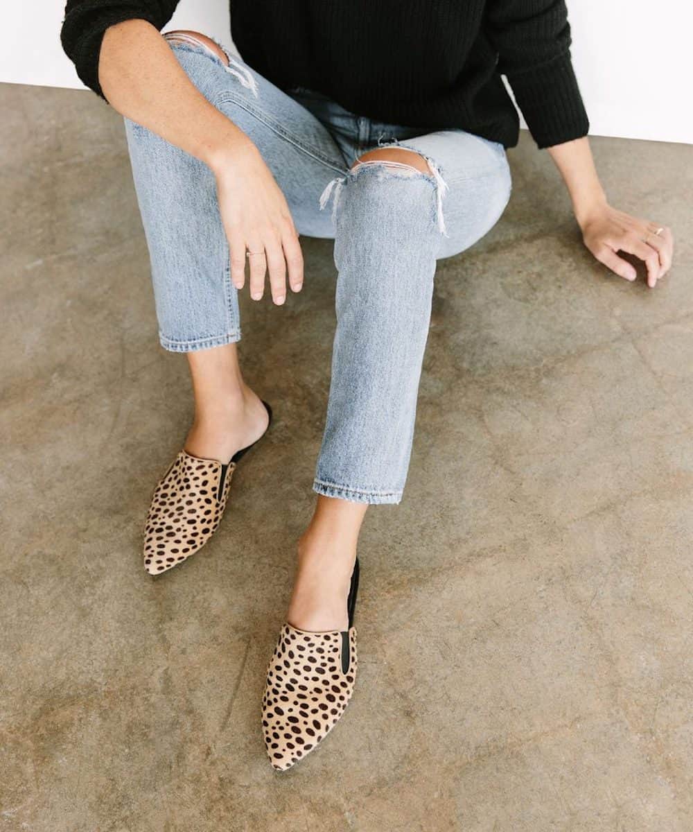 image of a woman sitting on a concrete floor wearing a black sweater, blue jeans, and cheetah print mules