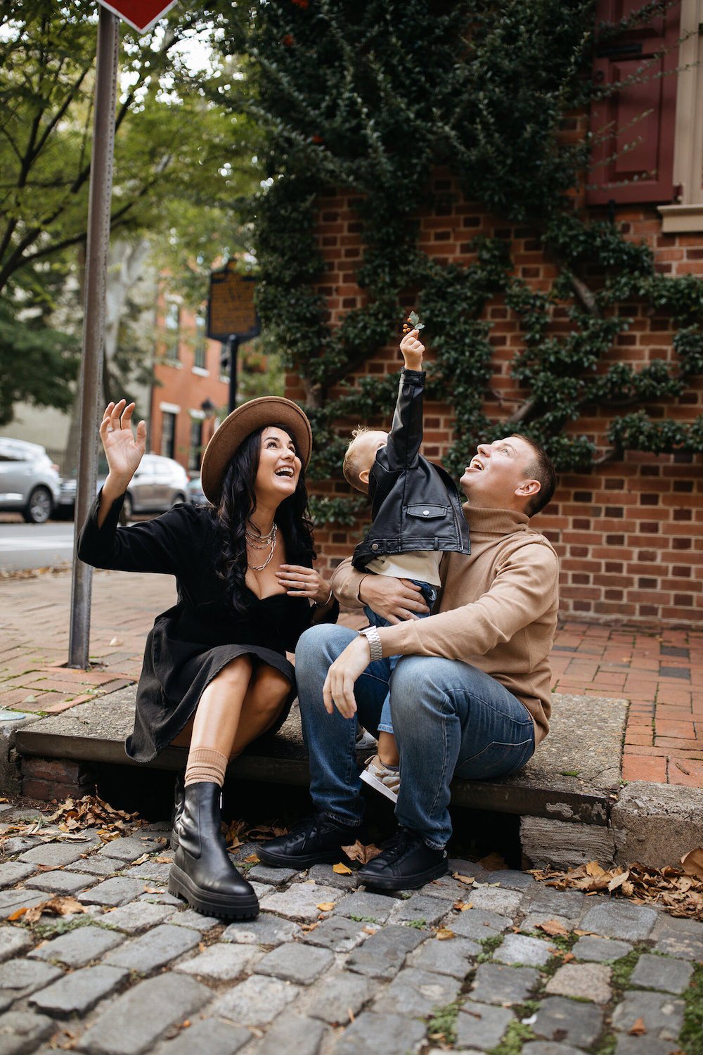 image of a family with a man, woman, and small child sitting on a sidewalk in front of a brick building in the fall wearing black, tan, and denim