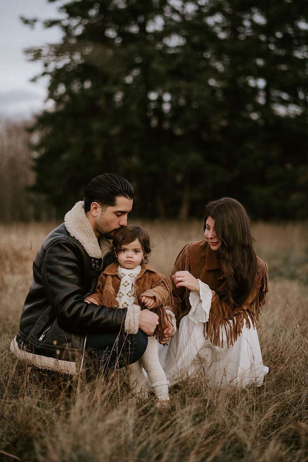 image of a family with a mom, dad, and baby in a field in the fall wearing brown suede fringe and black leather shearling jackets