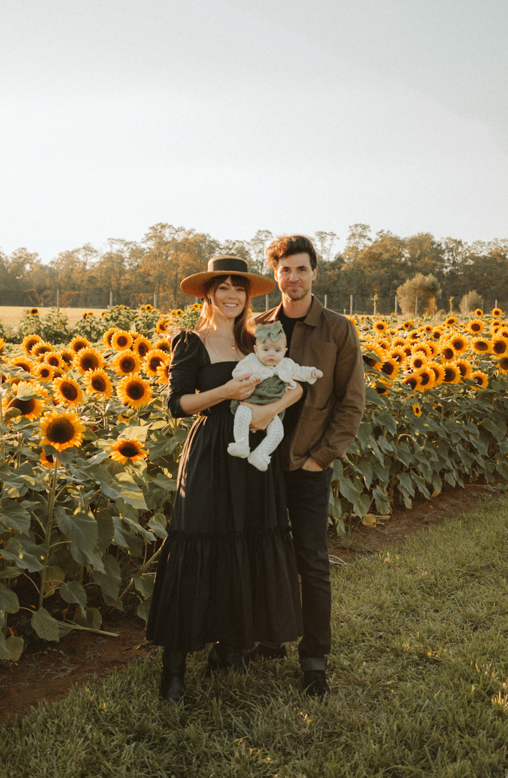 image of a family in a sunflower field, the mom wearing a black maxi sundress, the dad in a brown jacket and dark pants, and the baby in an ivory jumper