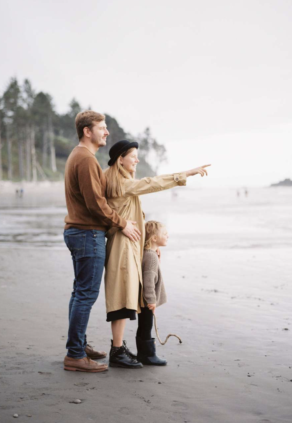 image of a family with a dad, mom, and small child wearing neutral clothing and boots standing on the beach looking out to the water in the fall