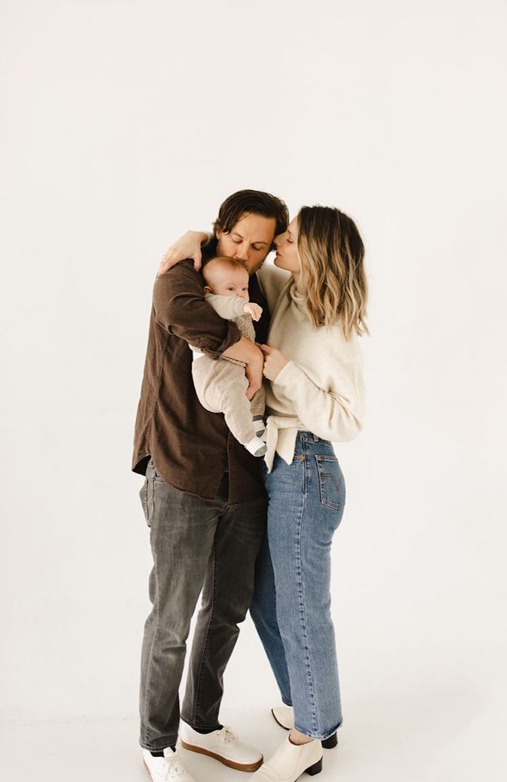 image of a family with a woman, man, and baby against a white wall for a family photoshoot, wearing jeans and casual tops