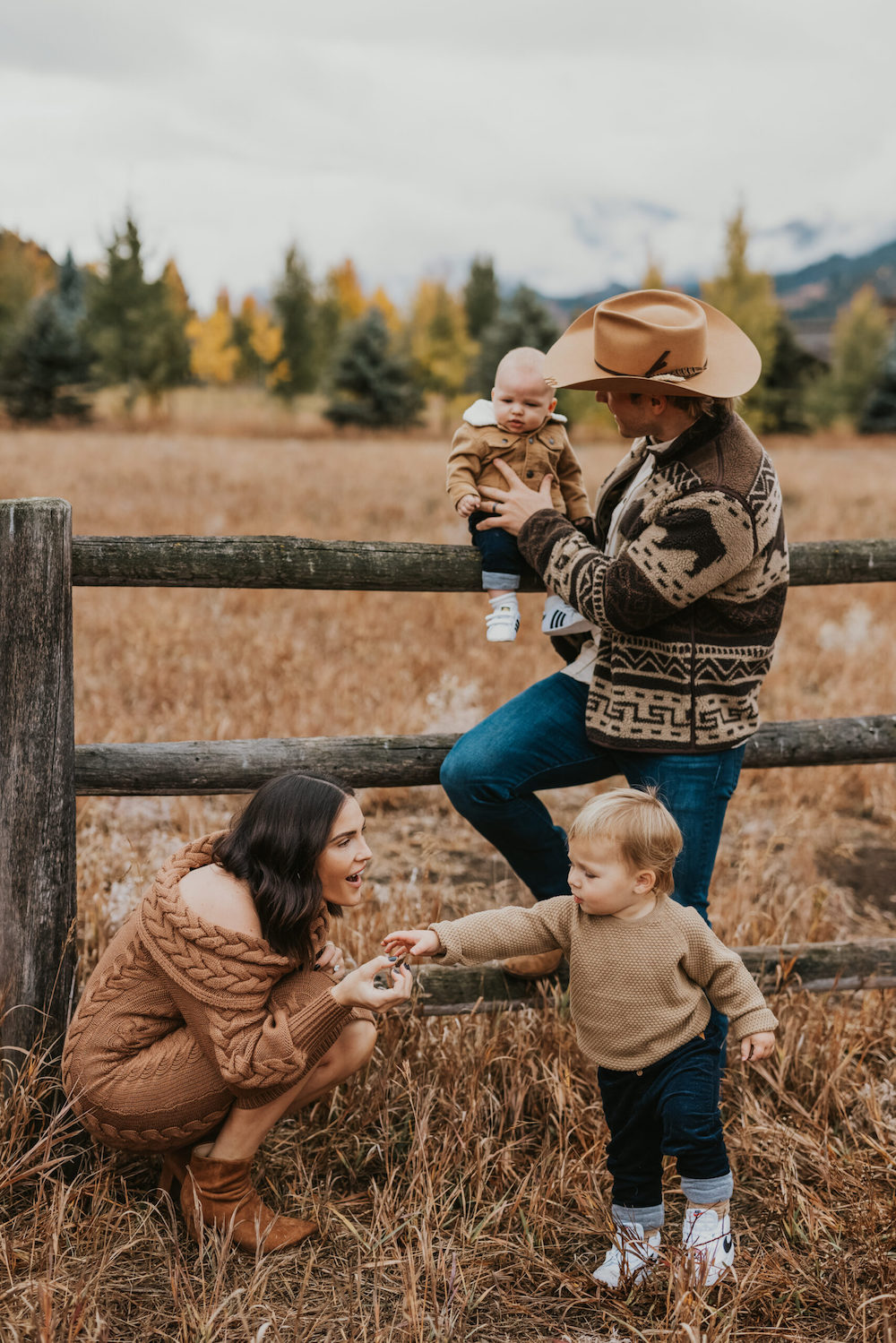 image of a family with a man, woman, toddler, and baby against a wooden fence in a field wearing neutral clothing and the man is in a cowboy hat