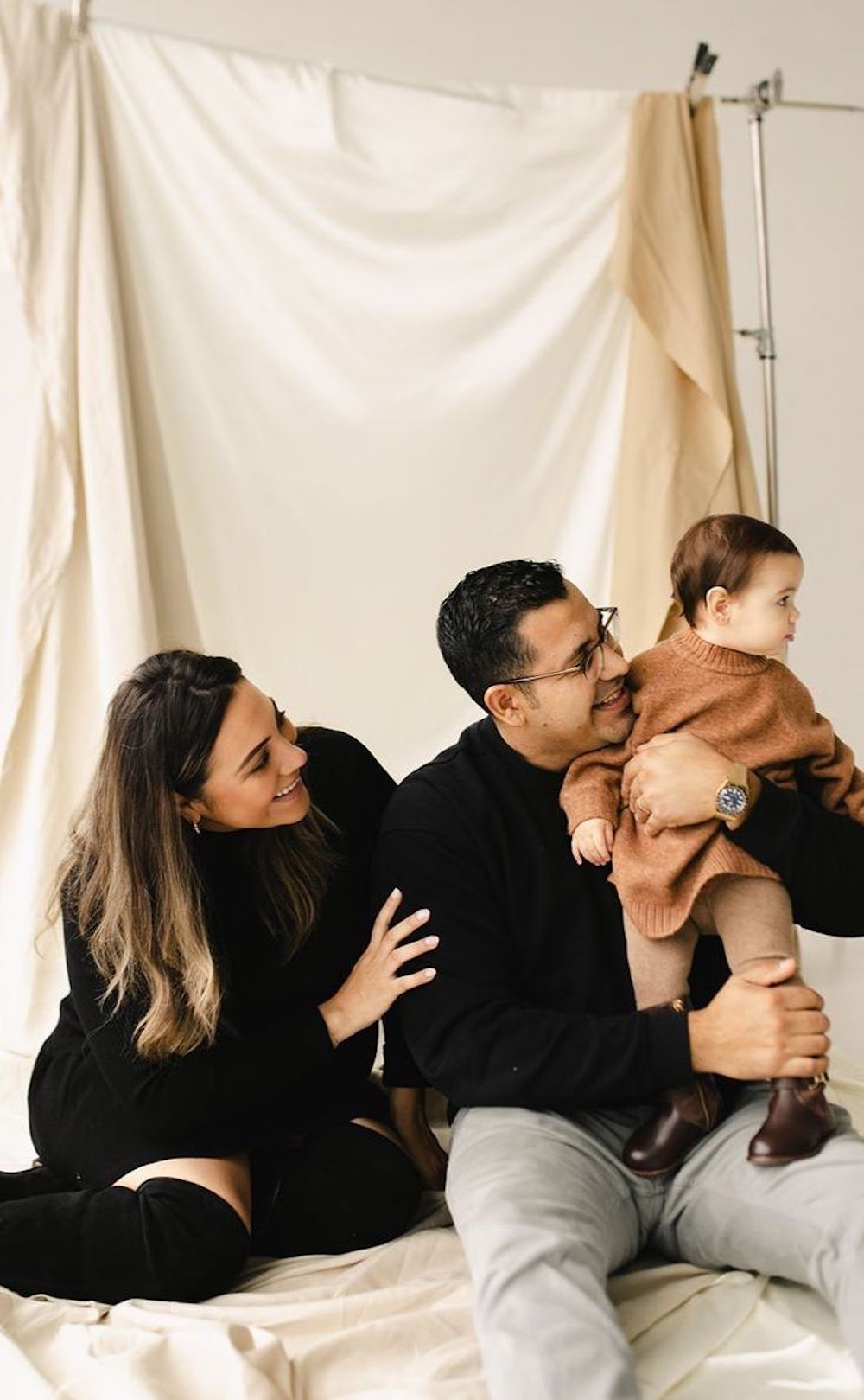 image of a family in a studio in front of a silk backdrop in black outfits with a toddler wearing a brown shirt and pants