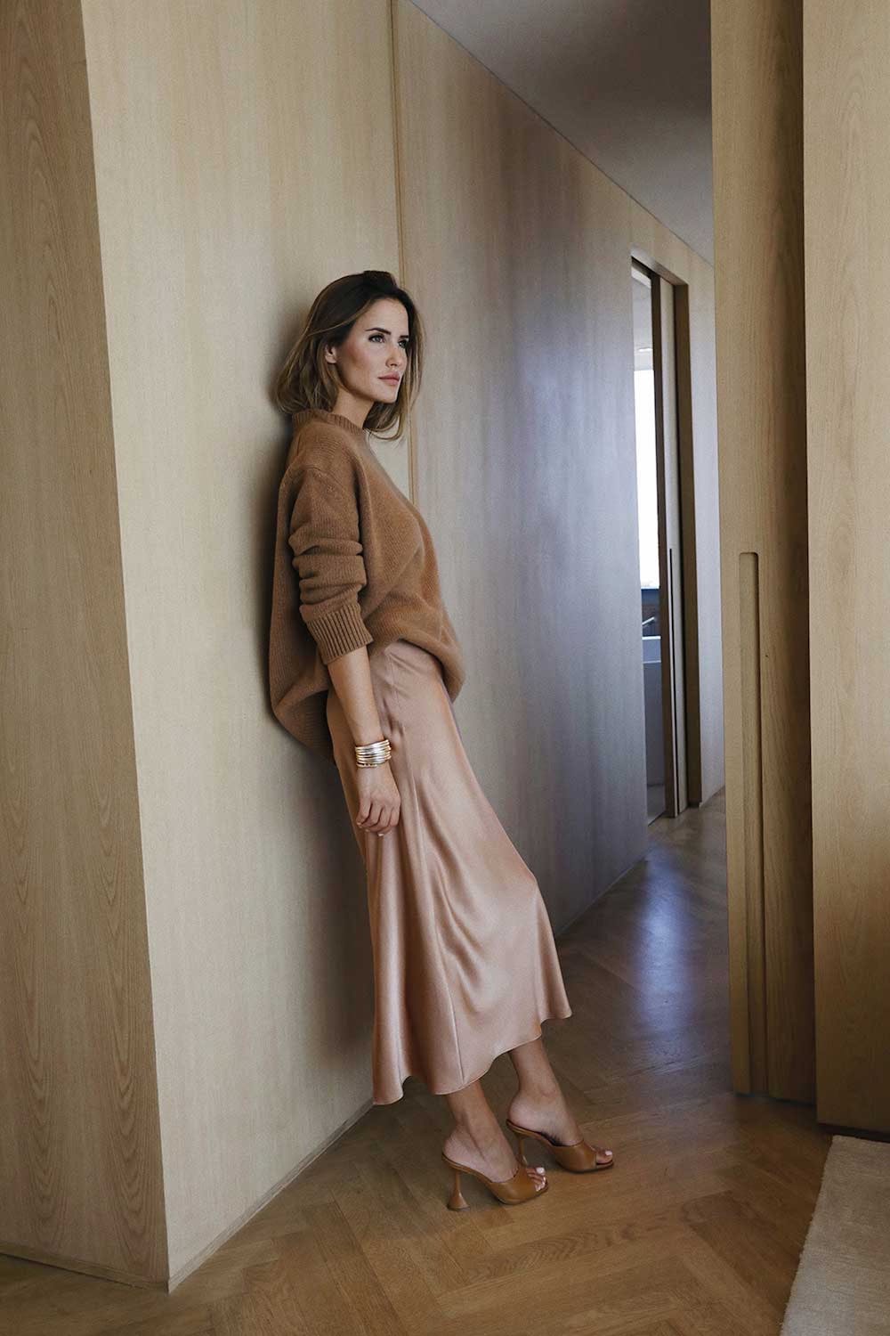 image of a woman leaning against a wall wearing a light brown silk midi dress with a brown sweater over top and brown heeled sandals