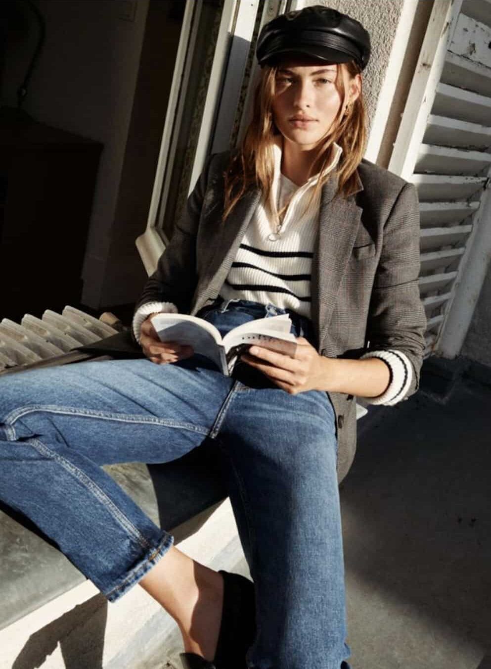 image of a woman in a blazer, striped knit sweater, and jeans leaning on a windowsill