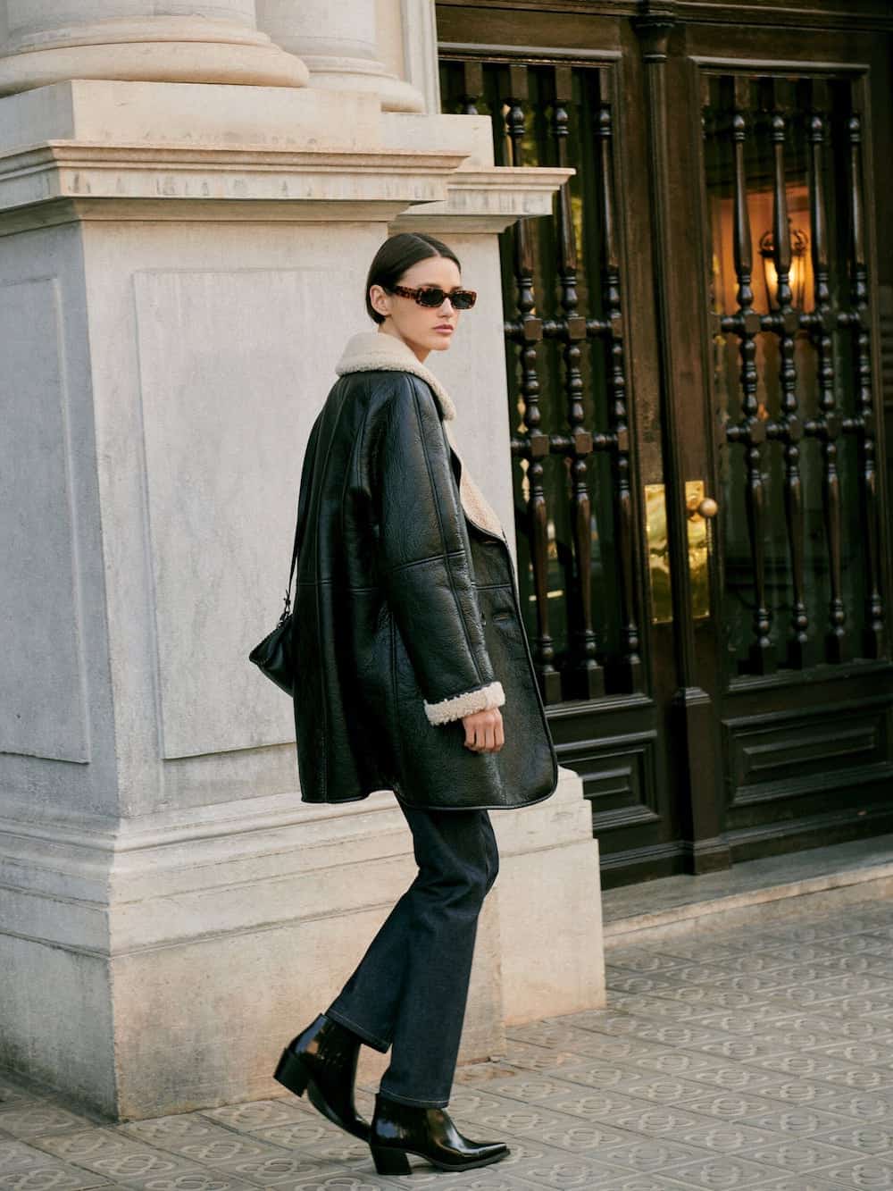 image of a woman walking down the sidewalk in a black leather and shearling jacket, dark jeans, and boots