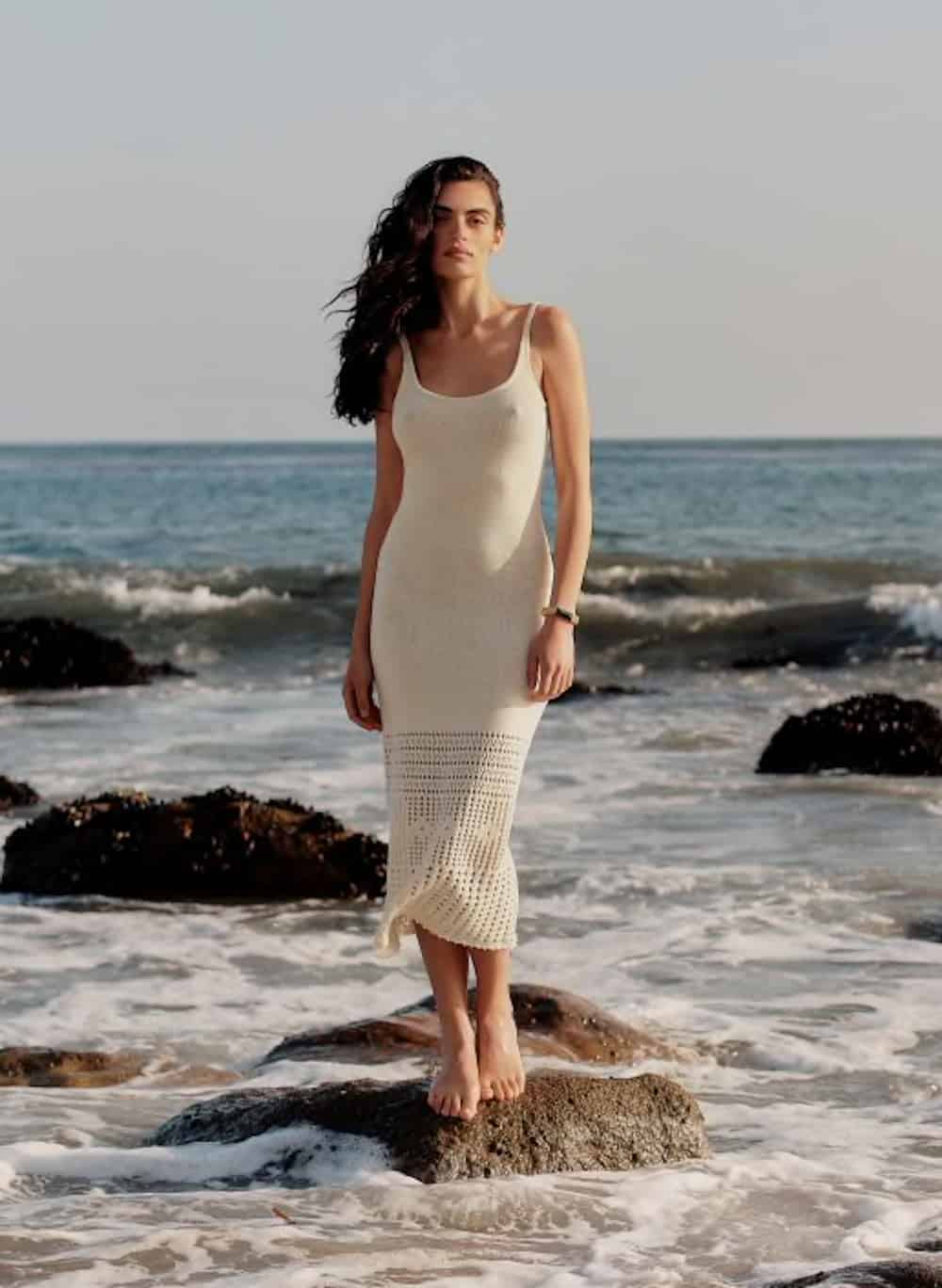 image of a woman in a long crochet knit ivory dress standing on a rock in the ocean