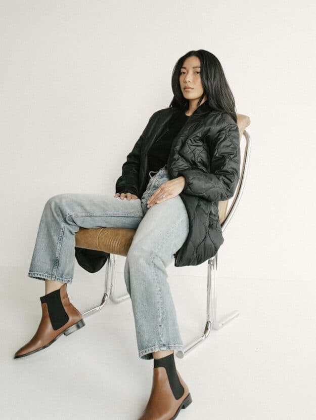 image of an asian woman sitting in a chair wearing faded denim jeans, brown boots, and a black puffer jacket